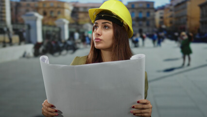 Hispanic woman architect in yellow helmet holding blueprints in st. peter's square, vatican, with...