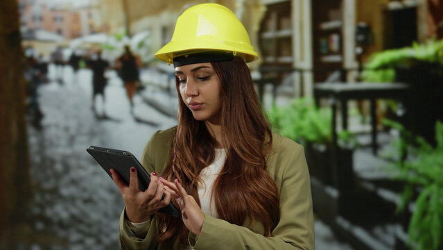 Woman wearing a yellow helmet and using a tablet, standing on an outdoor street, embodying a modern architect or engineer managing projects in an urban setting.