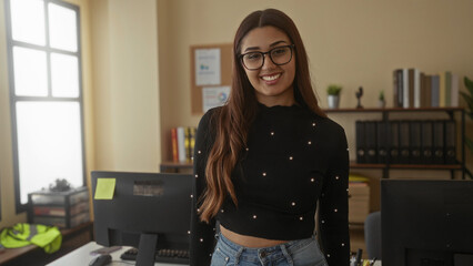 Woman standing confidently in a modern office environment with glasses and long hair, embodying professionalism and focus in an indoor work setting with natural light.