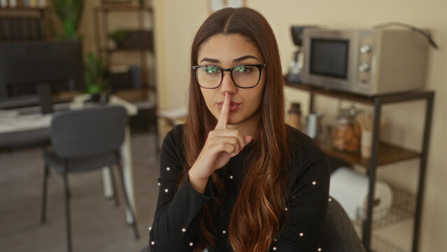 Hispanic woman making a silence gesture with finger on lips in an office interior, showcasing a modern work environment with a casual and thoughtful atmosphere.