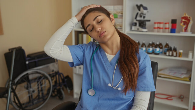 Hispanic woman nurse in blue scrubs looks tired in a hospital room with medical equipment and books on shelves beside a wheelchair, suggesting a busy workday.