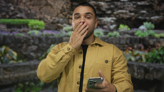 Young hispanic man holds smartphone and covers mouth while reading message on screen in lush green park; surprise.
