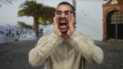 Man wearing glasses shouting with cupped hands by mouth in sunlit street; public announcement community excitement.