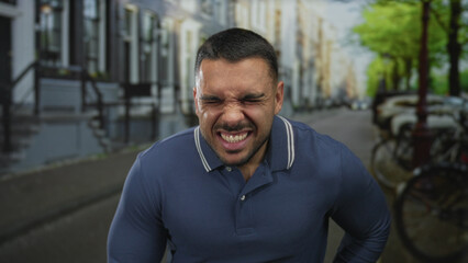 Naklejka premium Young hispanic man wearing a blue polo shirt gripping stomach and wincing on a crowded city street in daylight; pain.