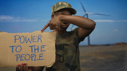 Young african american woman soldier in camouflage uniform points finger to cardboard sign in...