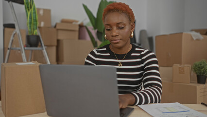 Young african american woman reads document at desk in building filled with boxes; serene determination.