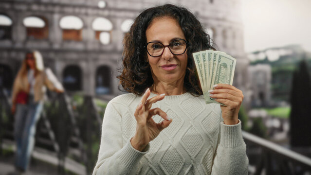 Fototapeta Woman holding american dollars near the roman coliseum makes an ok gesture, combining elements of travel and finance in an iconic italian outdoor city setting.