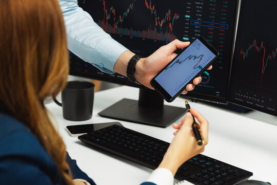 Cropped pointing hand with pen in businessman, checking with holding smartphone by woman investor online website program application with dynamic stock market at home office at back side. Infobahn.