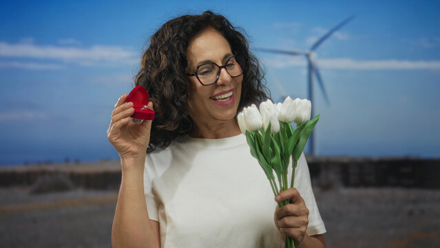 Hispanic woman joyfully holds a white flower bouquet and engagement ring near a windmill in a serene field backdrop, capturing a heartfelt outdoor proposal moment. - Powered by Adobe