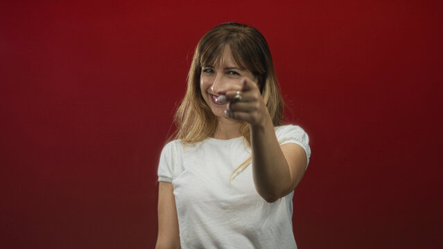Young caucasian woman points finger at camera in red studio, smiling with outstretched hand and direct gaze; confidence.