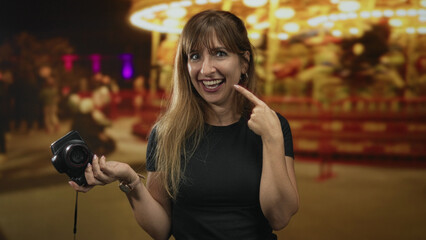 Woman holding camera points finger to cheek at a night street fair, smiling with teeth visible; joy celebration.