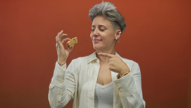 Woman pointing to crunchy oat granola bar in orange studio setting wearing white shirt; satisfaction hunger.
