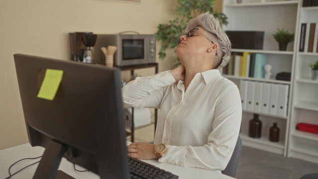 Woman in white shirt wearing glasses holding neck leaning back at office desk computer monitor; fatigue.