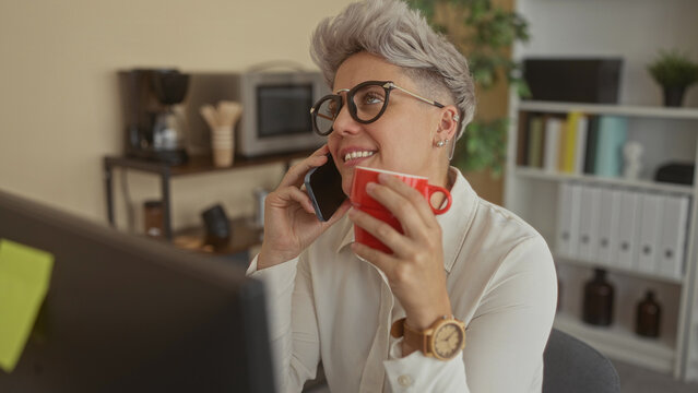 Woman holding smartphone to ear and red mug of coffee near computer monitor in bright office building; comfort connection productivity contentment.