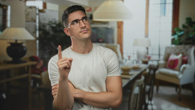 Man pointing finger with arm crossed in building living room, wearing glasses and white t shirt; thoughtful contemplation intent.