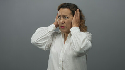 Hispanic middleaged woman covers ears with both hands in gray studio while appearing tense and concerned; frustration.