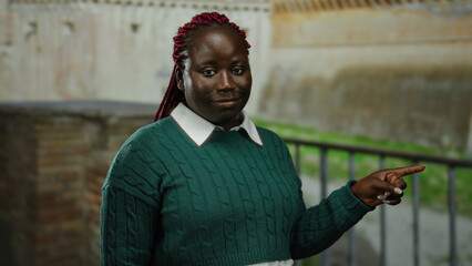 Woman with braids in green sweater pointing and smiling in roman ruins outdoor background in rome...