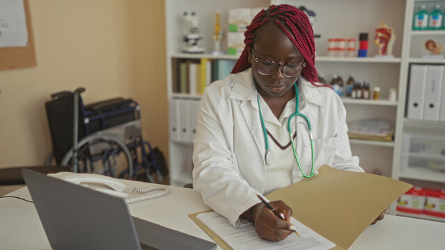 African american woman doctor writing notes in a hospital office, surrounded by medical equipment and documents, highlighting professional healthcare work indoors.