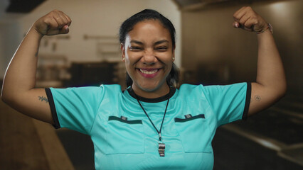 Woman referee wearing turquoise shirt and whistle flexing arms in kitchen indoors; strength empowerment confidence.