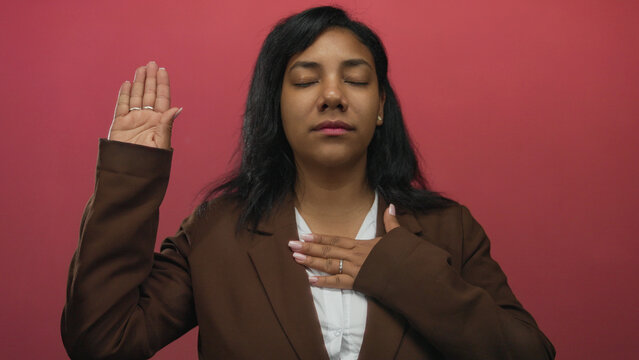 African american woman raises hand and touches chest with eyes closed in pink studio; pledge sincerity.