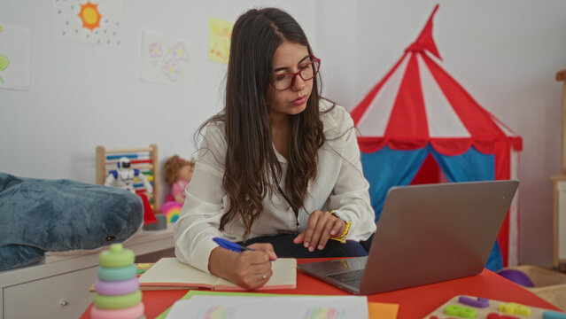 Young woman teacher in a colorful kindergarten taking notes with a laptop on a table surrounded by educational toys and vibrant decor indoors during a lesson preparation session.