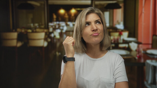 Seated young blonde woman covers mouth with hand while yawning at a wooden table in a restaurant; boredom.