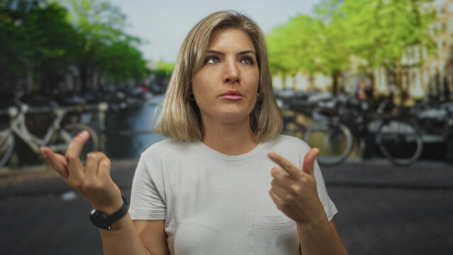 Young blonde woman wearing white t shirt and black wristwatch pointing fingers upward on street in amsterdam canal district; uncertainty.