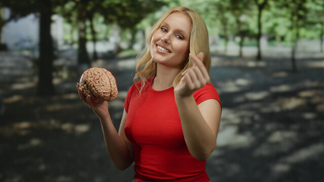 Woman in a park holding a brain while making a heart gesture, surrounded by green trees on a sunny day indicating a sense of curiosity and love for science and nature.