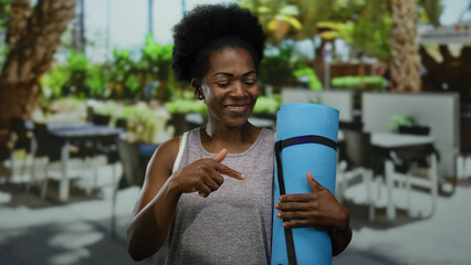 Woman holding yoga mat on a city terrace, surrounded by outdoor restaurant seating, showcasing a blend of fitness and urban life during a warm day.