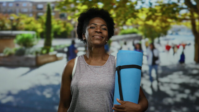 Woman holding yoga mat outdoors on city street smiling in athletic gear surrounded by blurred urban environment with trees and buildings in background. - Powered by Adobe