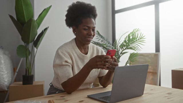 Woman using smartphone in bright new apartment with laptop on table surrounded by moving boxes and plants suggesting modern lifestyle transition and connection.