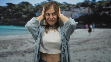 Woman covers ears with both hands on a sandy beach by the rocky coast, posing near shoreline cliffs; annoyance.