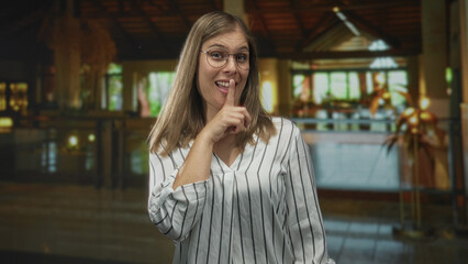 Young blonde woman points finger to side in building lobby wearing striped blouse and glasses, visible forefinger gesture; playful teasing.