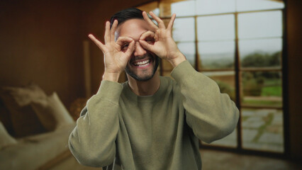 Playful young hispanic man indoors makes glasses gesture with hands in a cozy rustic mediterranean house, smiling joyfully by window with nature view in the background.