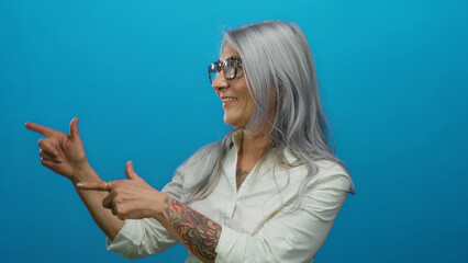 Grey-haired senior woman with tattoo smiles and points against a vibrant blue wall, wearing glasses and a white shirt, conveying confidence and style.