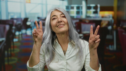 Senior woman with grey hair making a peace sign in an indoor restaurant setting, exuding positivity and confidence amidst colorful decor and modern interior design.
