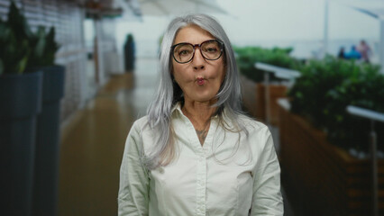 Senior woman with grey hair makes a humorous face on a bustling outdoor street, surrounded by blurred people and greenery, wearing glasses and a white shirt.