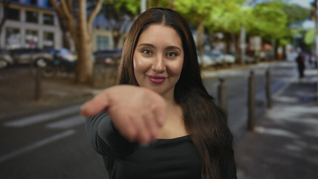 Young hispanic woman reaches out palm forward with a smile on a sunlit street; friendly invitation warmth.