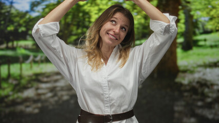 Naklejka premium Woman smiling and raising hands above head in forest path, wearing white shirt and belt while facing camera; joy playfulness energy.