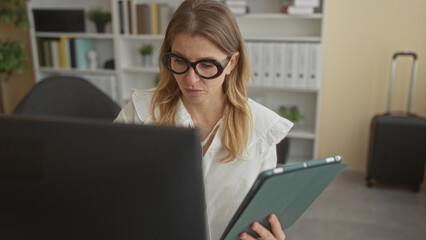 Woman wearing round glasses holds a tablet and looks at a laptop screen in an office building with suitcase and shelves visible; concentration productivity efficiency.