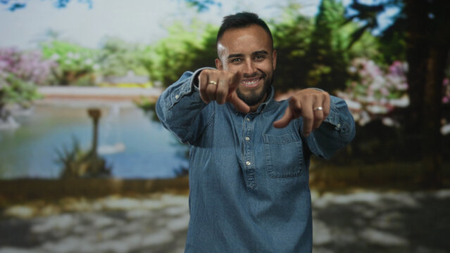 Man points finger at camera in park, smiling with bare hands and rings visible, wearing denim shirt and beard visible; confidence success.