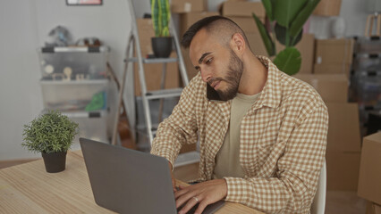 Man talking on phone at home at a table with laptop and hand on keyboard, packed boxes and ladder visible; moving productivity.