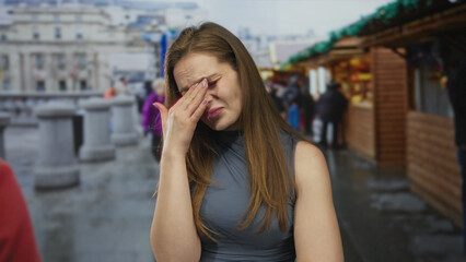 Young caucasian woman wiping tears with hand over eye in a busy street market lined with wooden souvenir stalls; sadness.
