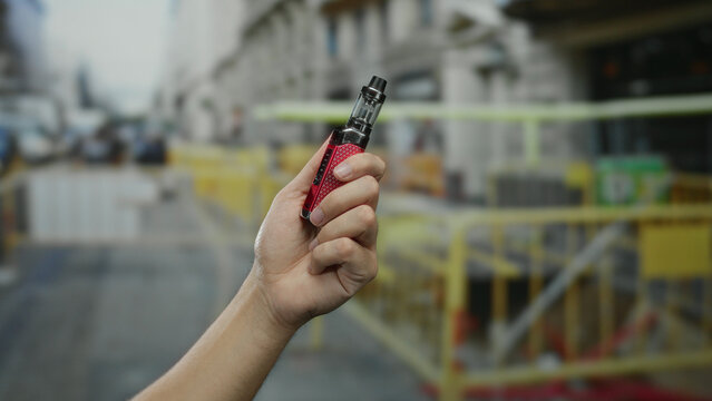 Man holding a vape outdoors at a construction site, showcasing the device in a street setting surrounded by urban structures.