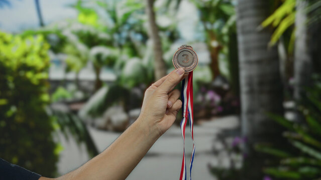 Man hand holding bronze medal with ribbon in vibrant park setting showcasing achievement against lush outdoor greenery giving a sense of success and victory celebration.