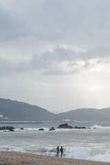 Zwei Personen stehen am Rand eines sandigen Strandes vor einer rauen See, im Hintergrund sind H&uuml;gel, Industrieanlagen und ein bew&ouml;lkter Himmel mit diffusem Licht zu sehen. Die Atmosph&auml;re wirkt ruhig u