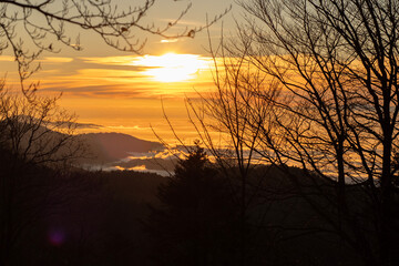 Die Sonne geht hinter bewaldeten H&uuml;geln im Schwarzwald unter, der Himmel leuchtet in warmen Orange- und Gelbt&ouml;nen, Nebelschwaden liegen im Tal