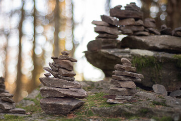 Aufeinander gestapelte Steine auf einem moosbewachsenen Felsen im Wald. Die Sonne scheint durch die B&auml;ume im Hintergrund, das Bild wirkt ruhig, meditativ und naturverbunden