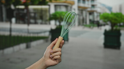 Man holding whisk on city street with blurred urban background, capturing a unique blend of everyday kitchen tool in an unexpected outdoor urban setting.