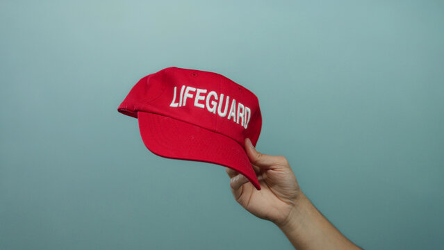 Man holding red lifeguard cap against blue wall showcasing safety and aquatic theme. - Powered by Adobe